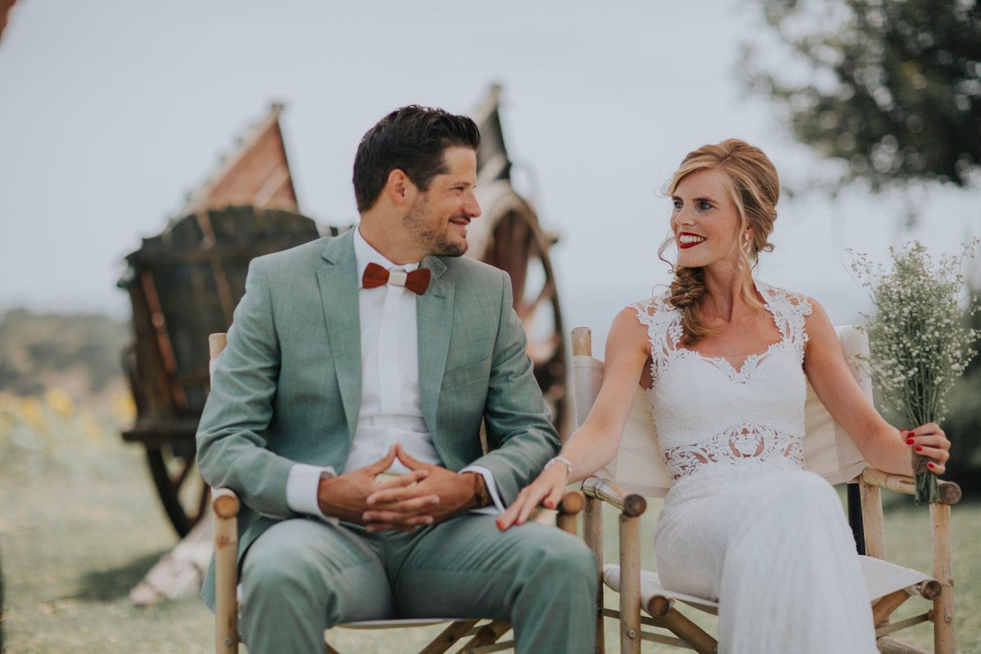 A bride and groom share a candid moment while sitting in vintage chairs during their outdoor wedding ceremony.