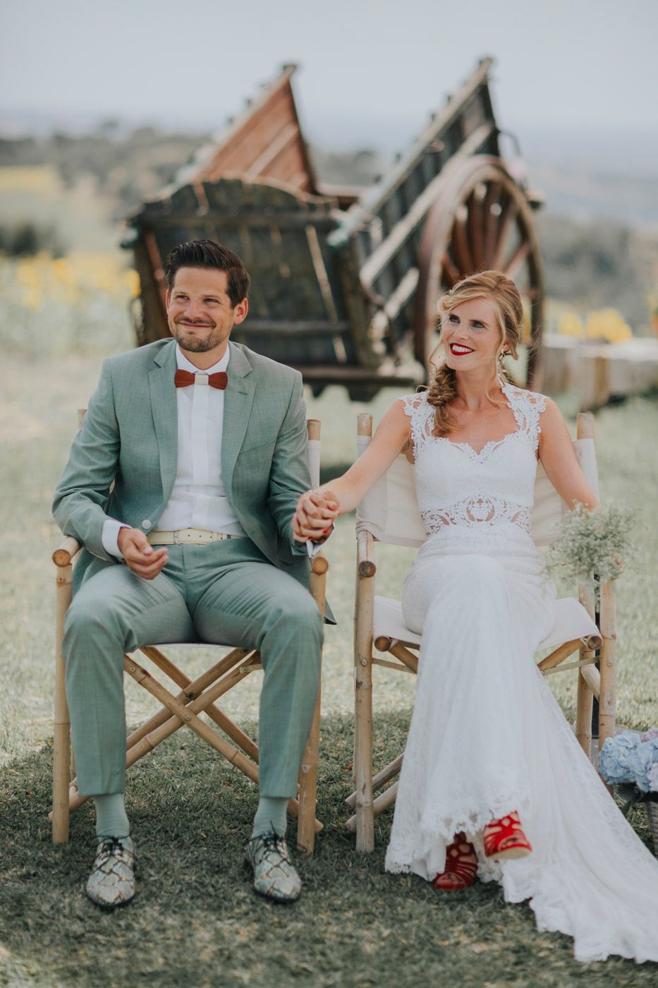 A bride and groom sit in wooden chairs outdoors with rustic structures in the background.