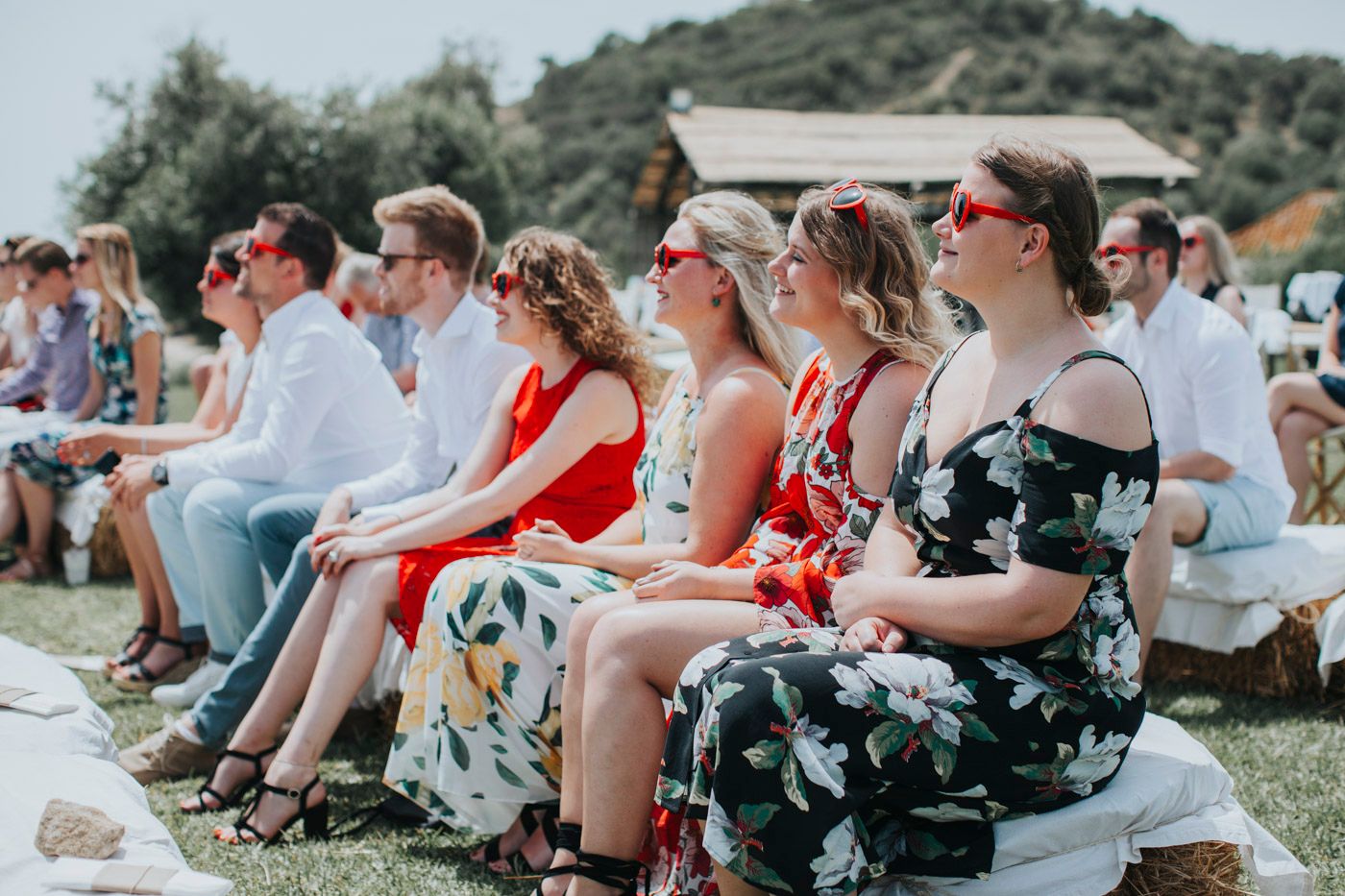 Wedding guests sitting outdoors in summery attire watch ceremony at scenic hillside location.