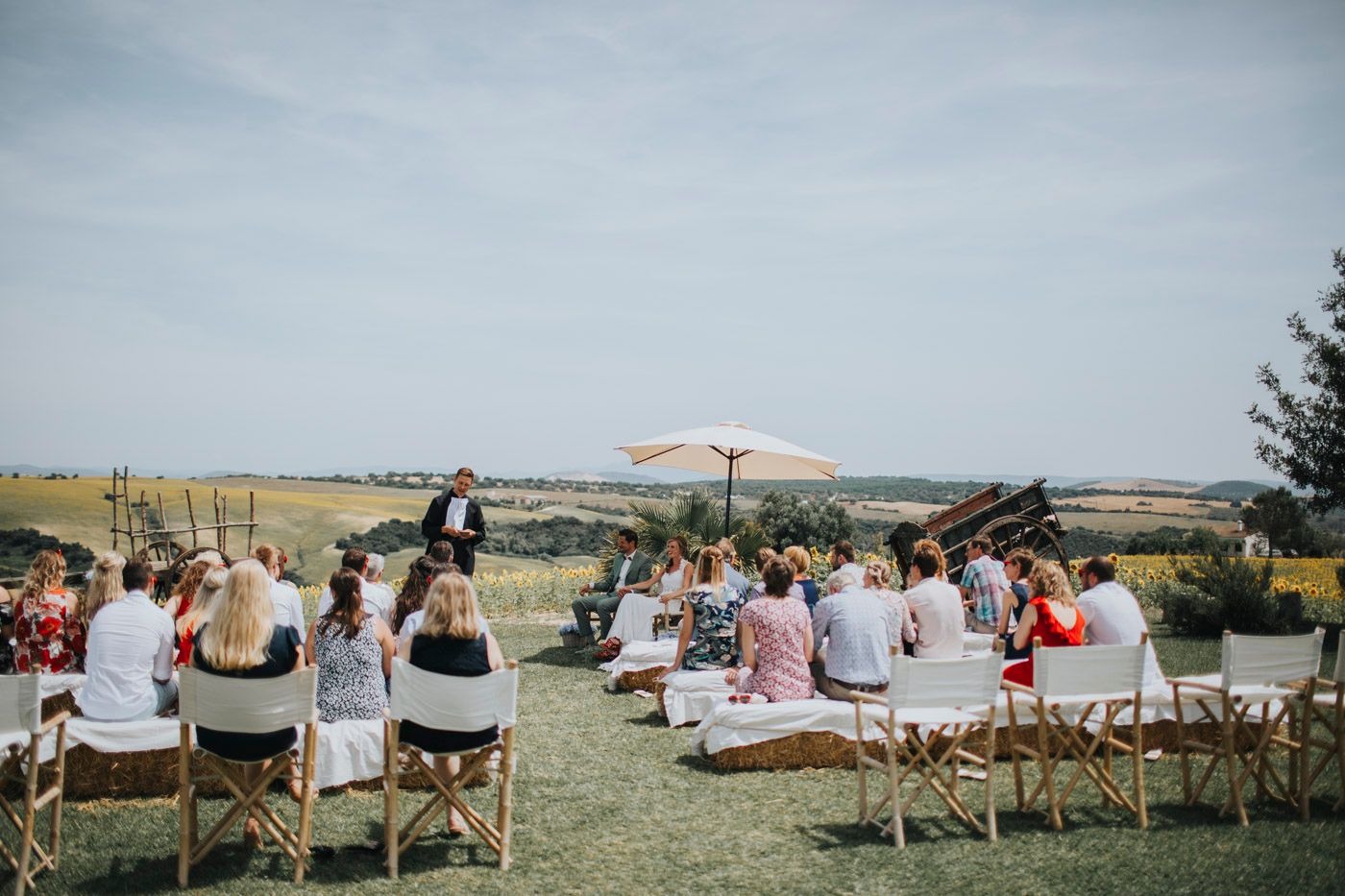 Outdoor wedding ceremony guests seated on folding chairs and blankets under a cloudy sky in a rural setting.