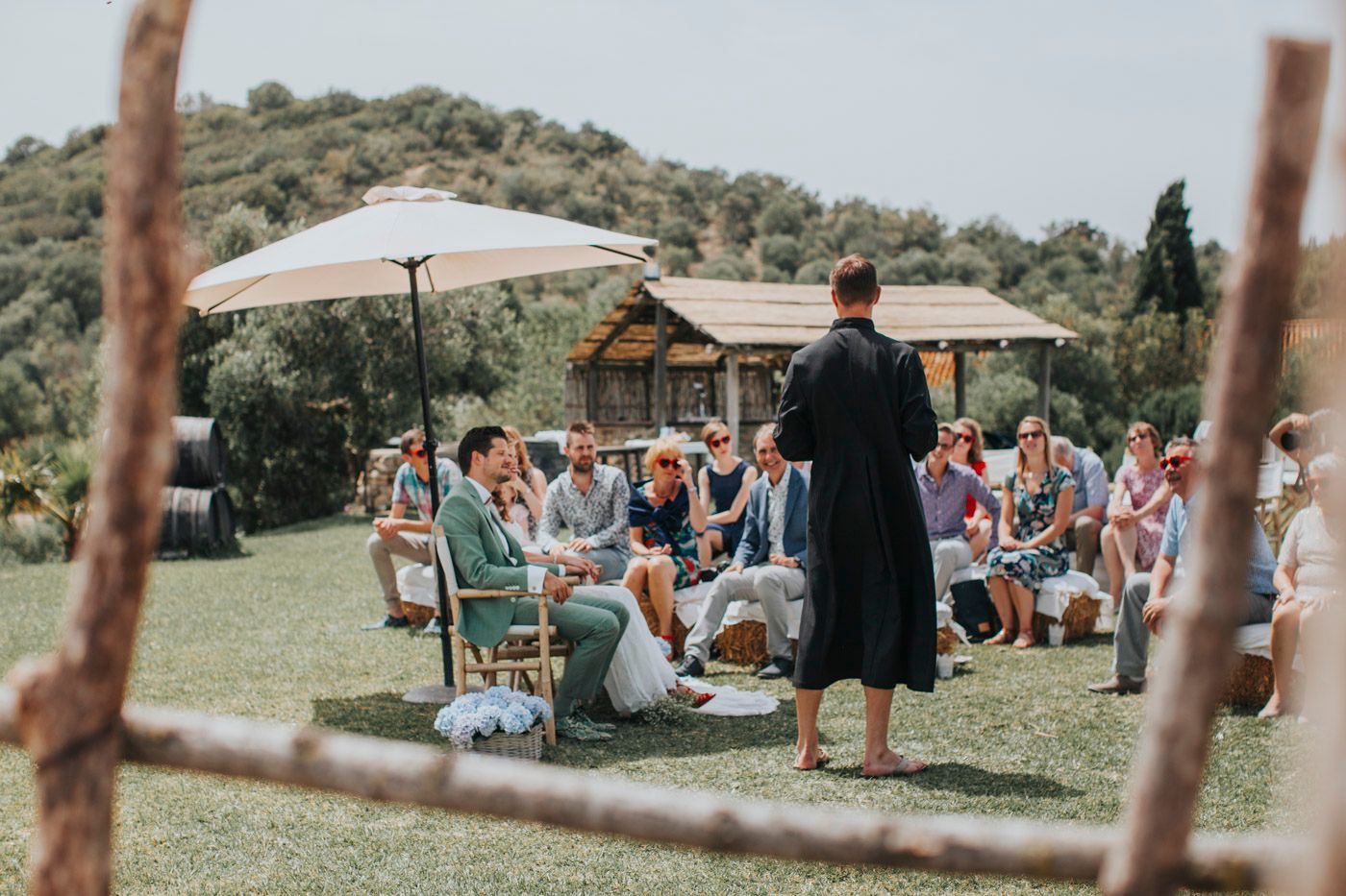 A small outdoor wedding ceremony in a rustic setting with white umbrellas and wooden chairs on a green hillside.