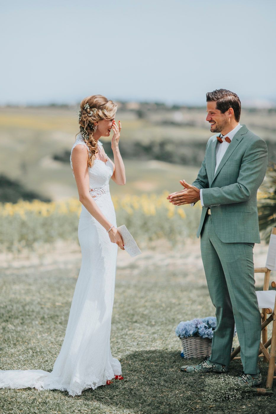 A bride in white lace gown and groom in mint green suit share an emotional moment in an outdoor wedding ceremony.