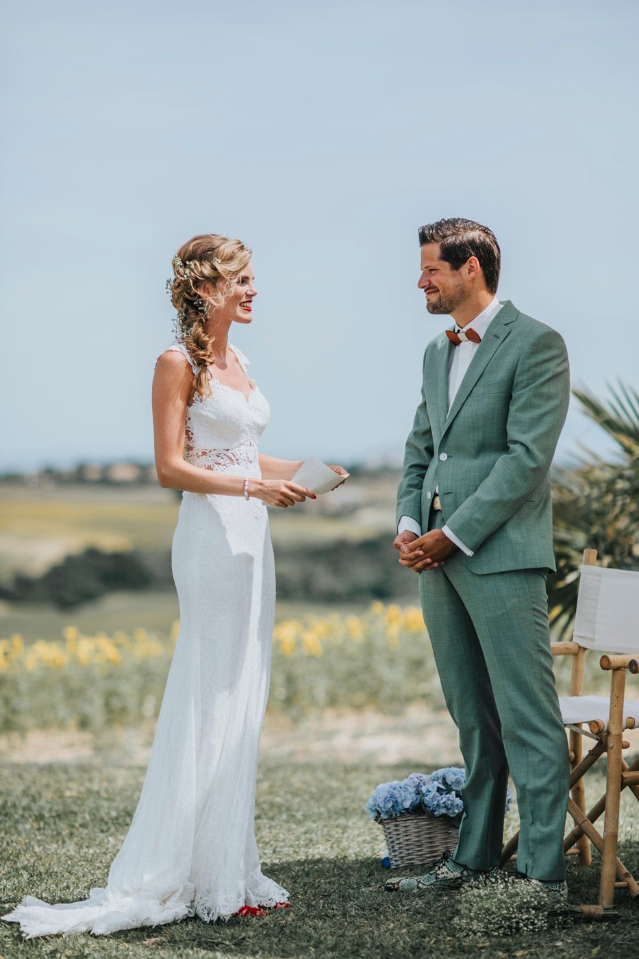 A wedding couple exchanges vows outdoors with a sage green suit and flowing white dress against a scenic backdrop.