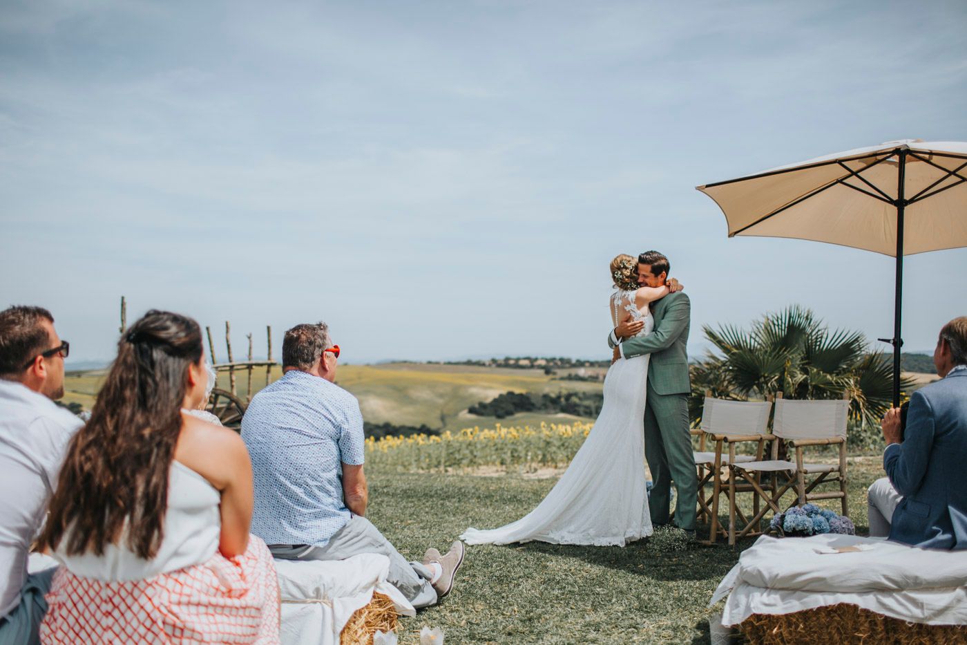 An intimate outdoor wedding ceremony takes place on a grassy hilltop under a beige umbrella with guests seated on chairs.