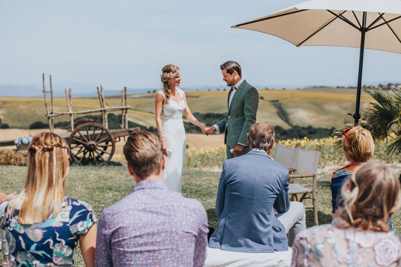 An intimate outdoor wedding ceremony under a white umbrella with guests seated on a grassy hillside overlooking the countryside.