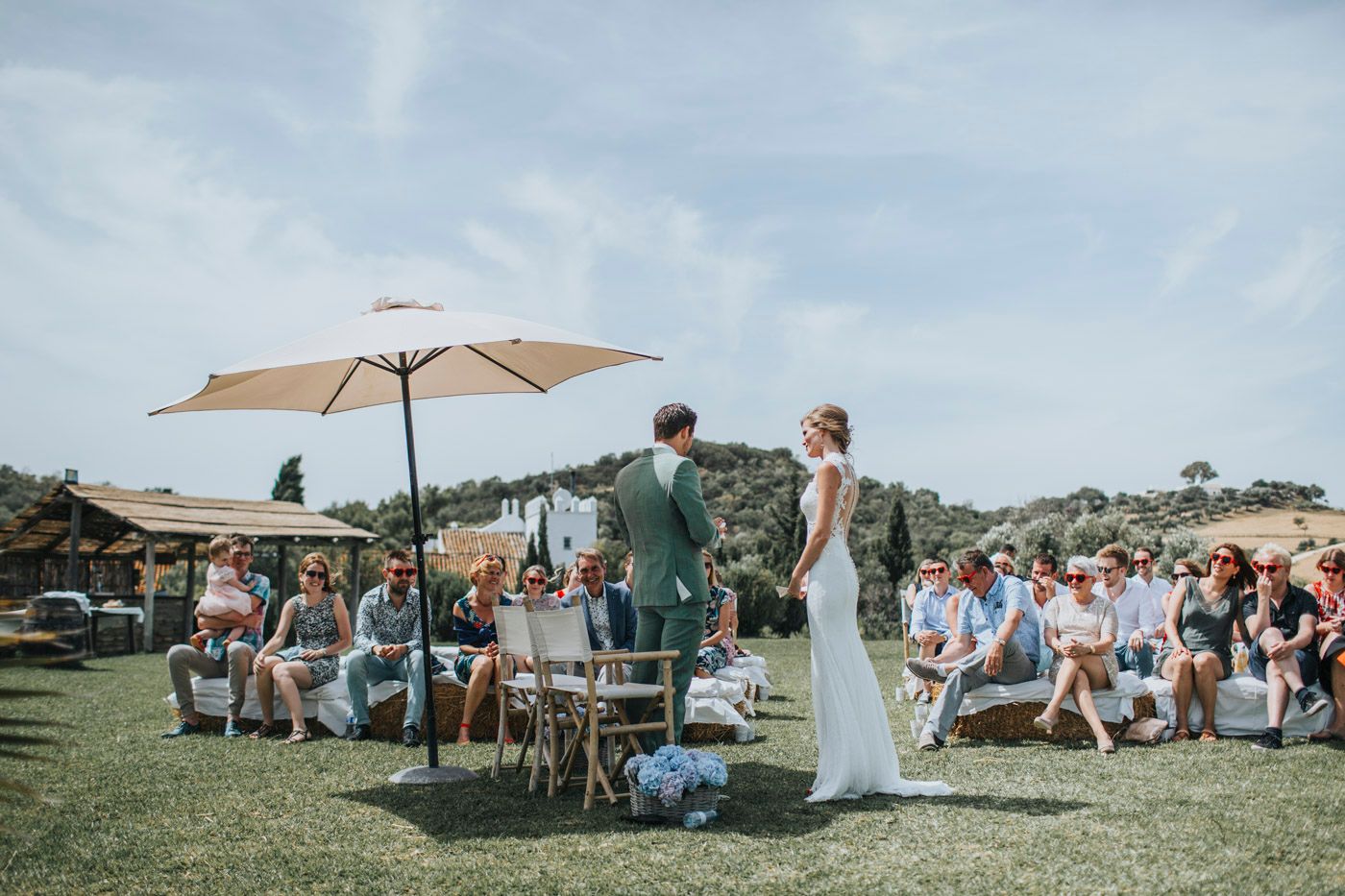 An outdoor wedding ceremony on a grassy lawn with guests seated under a sunny sky with a beige umbrella stand.