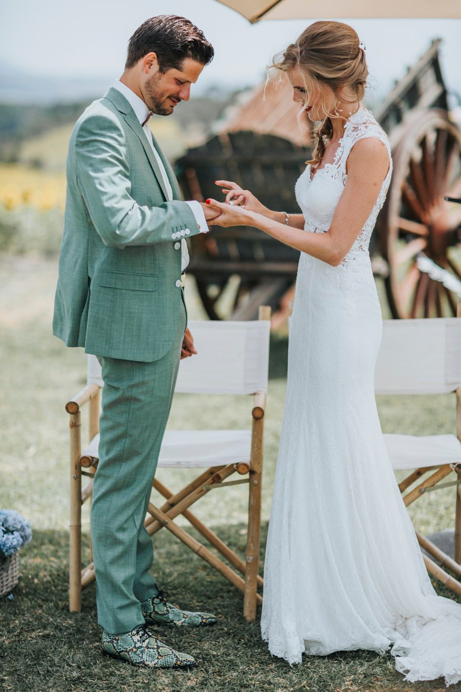 A couple exchanges rings during an outdoor wedding ceremony with the bride in white lace and groom in mint green suit.