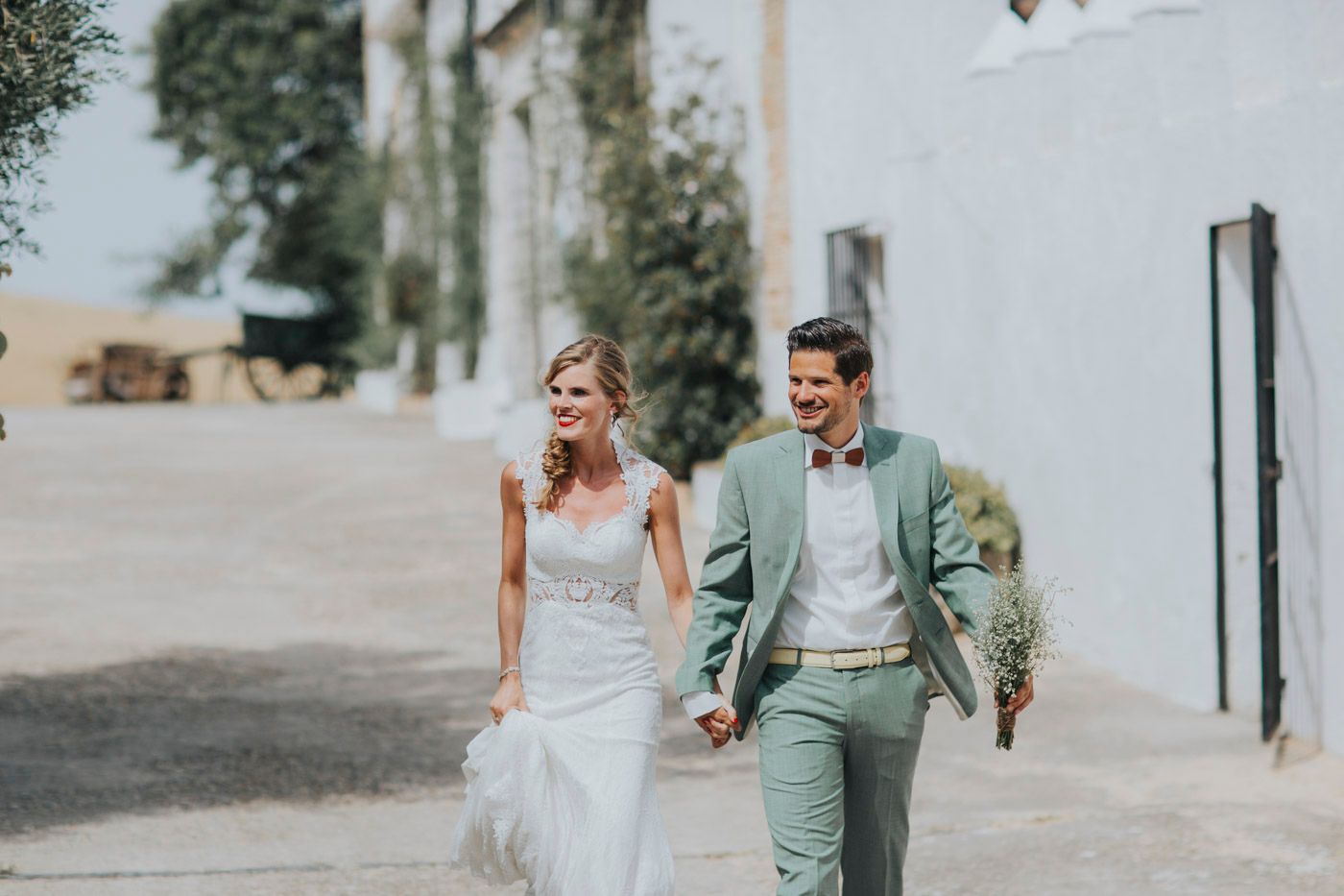 A couple in wedding attire walk and laugh together along a rustic building wall on their special day.