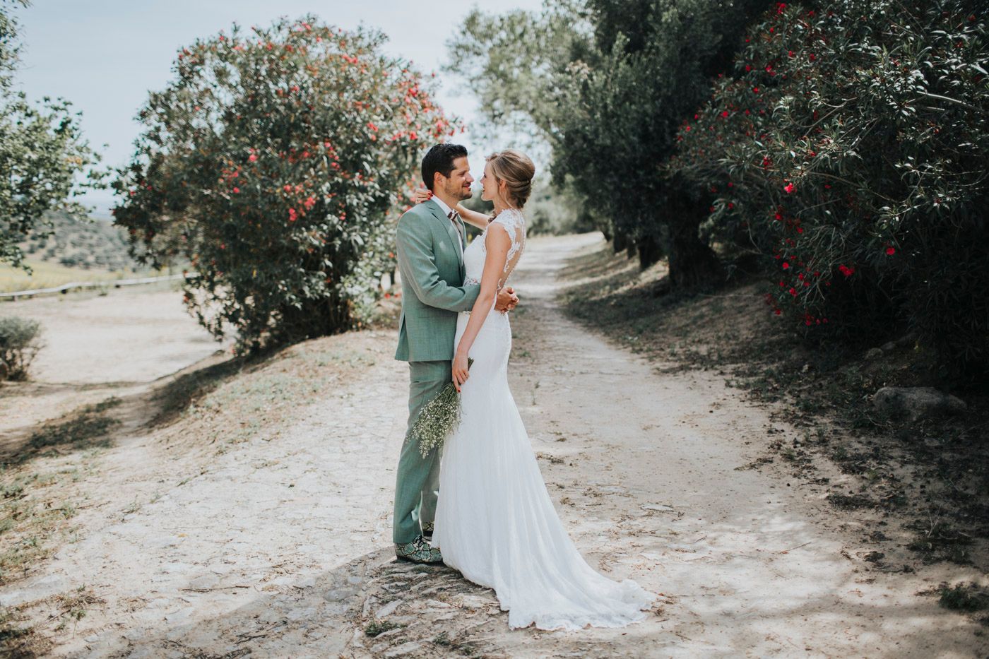 A couple embraces on a sunlit dirt path surrounded by flowering trees and greenery on their wedding day.
