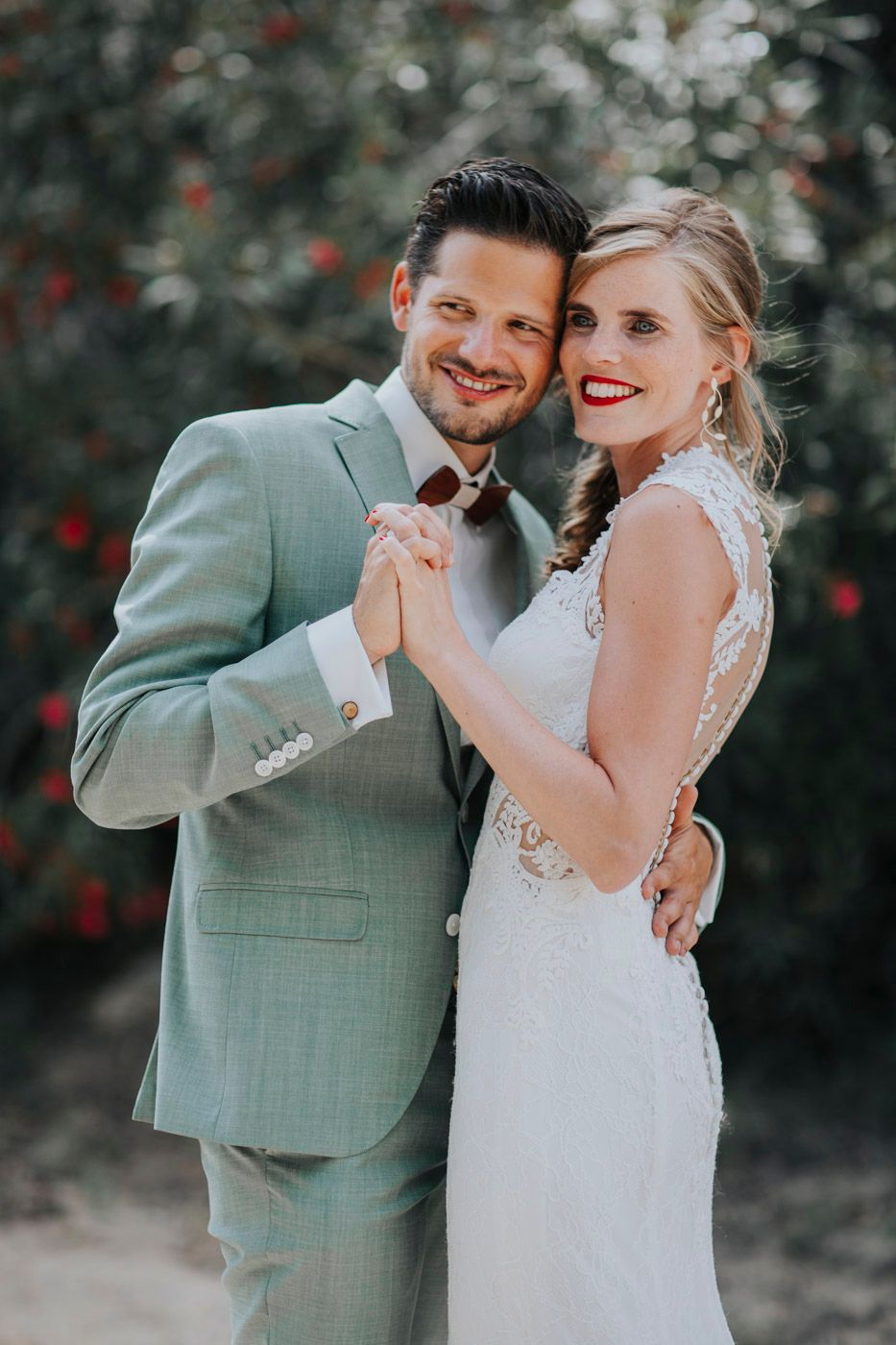A romantic wedding portrait of a couple in a sage green suit and white lace dress holding hands and smiling.