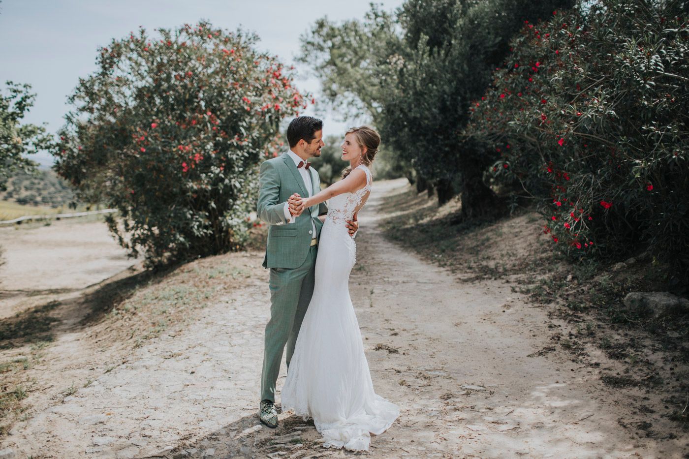 A couple shares a romantic moment on a dirt path surrounded by blooming red flowers and olive trees.