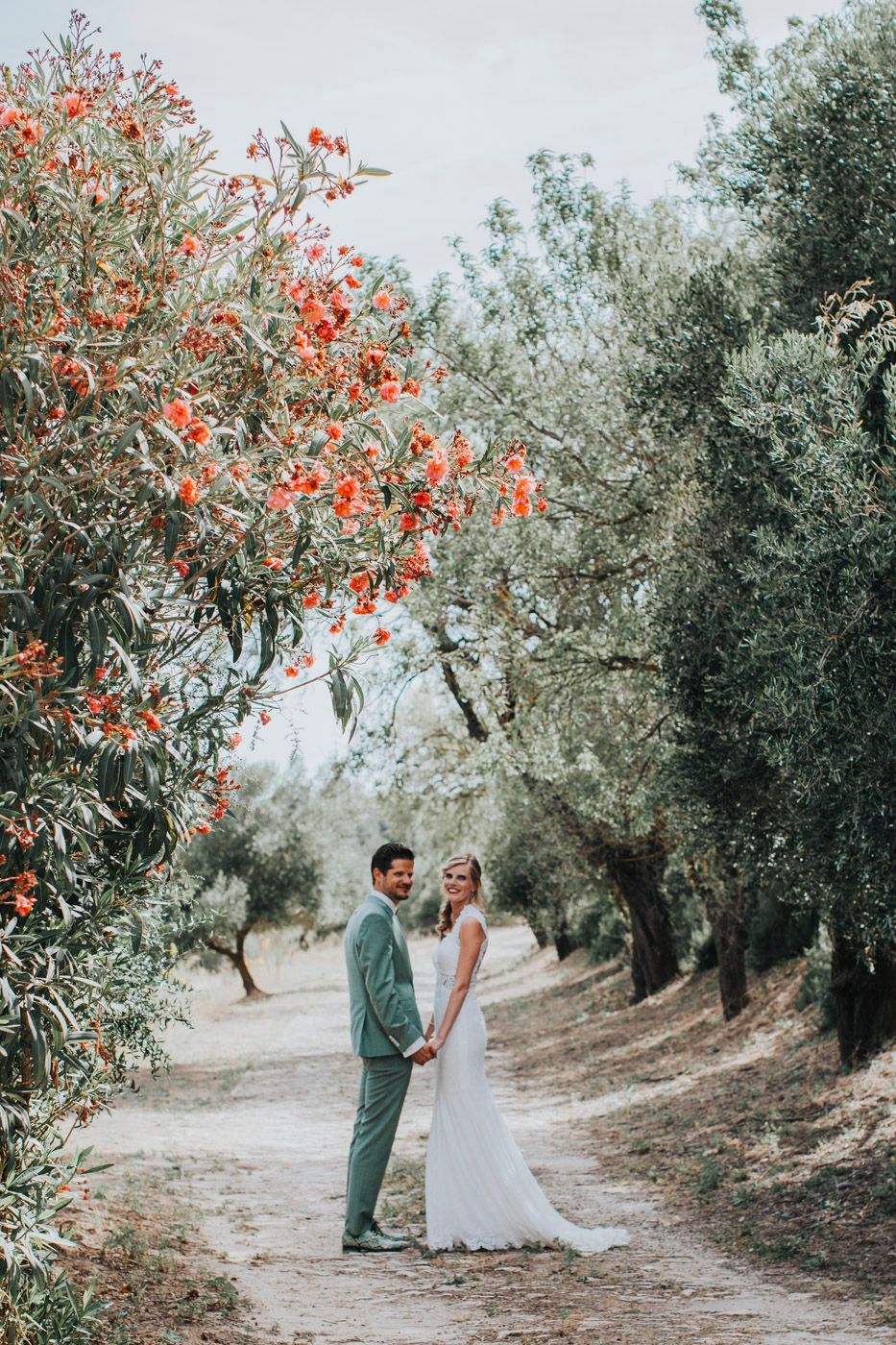 A newlywed couple poses on a path surrounded by olive trees and orange blossoms in a dreamy outdoor setting.