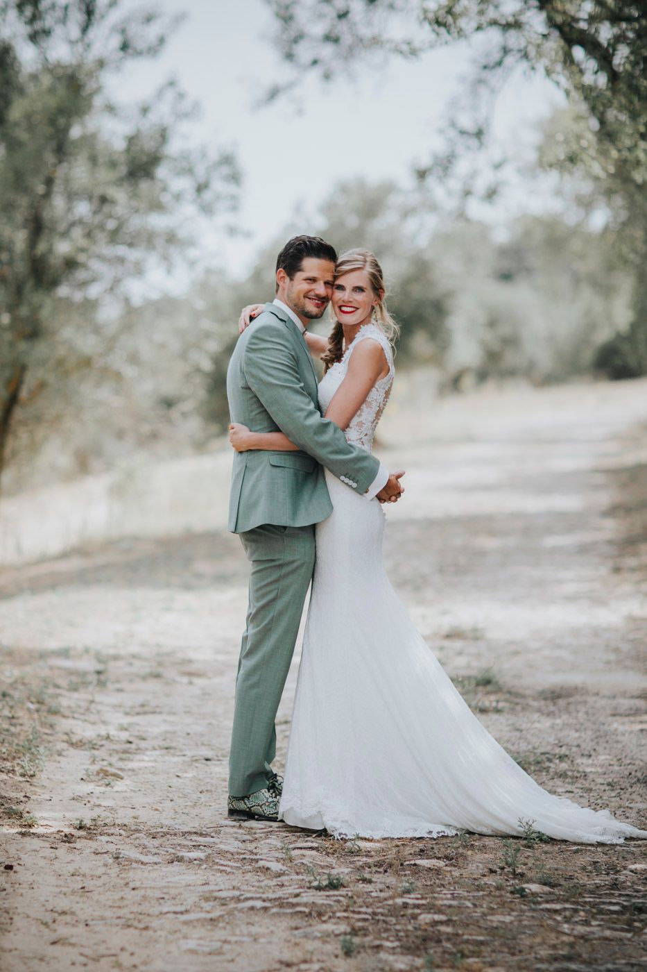 Happy couple embraces on a dirt path surrounded by trees during outdoor wedding photoshoot.