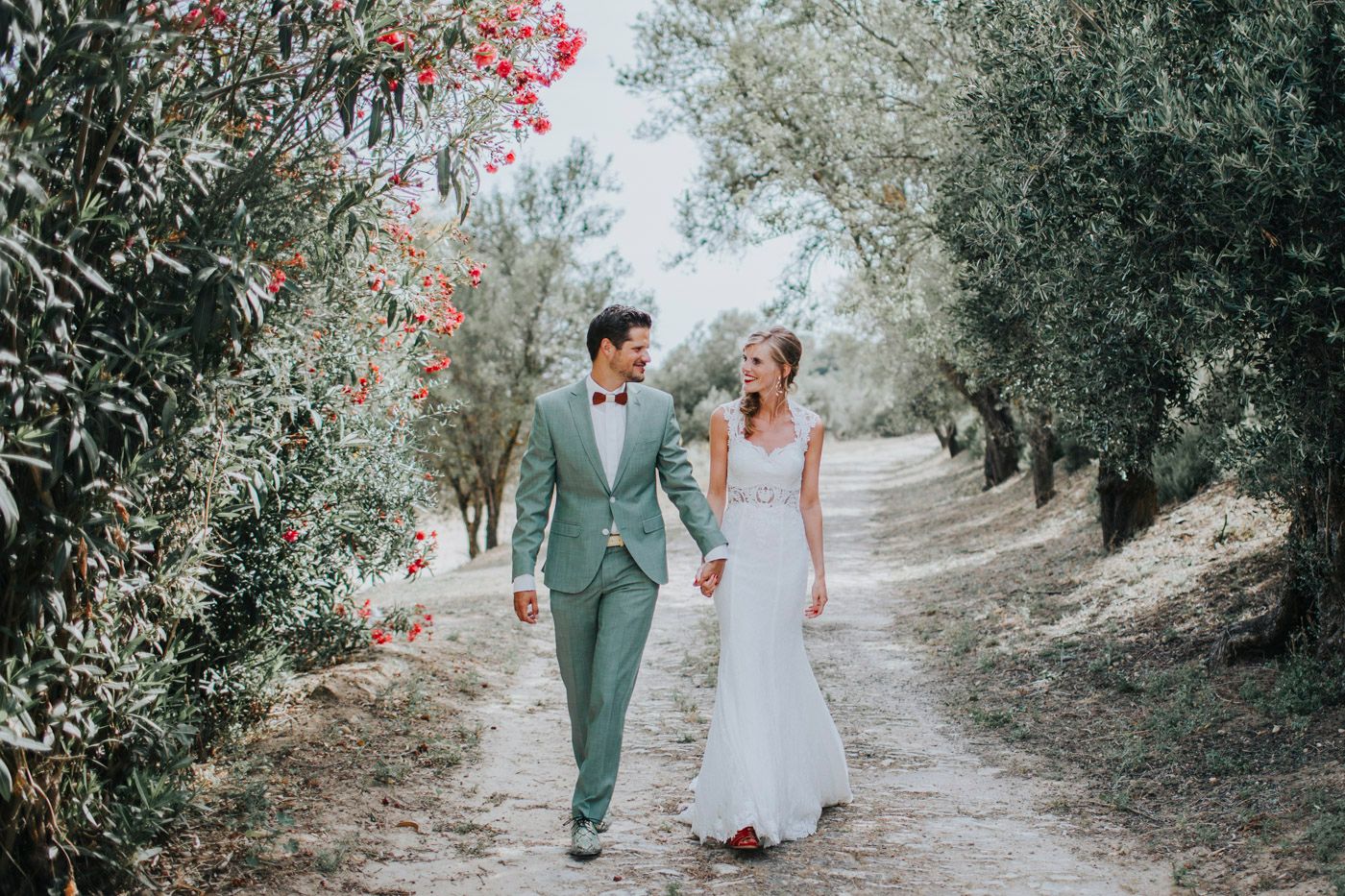 A romantic wedding couple walks hand in hand down a tree-lined path with pink flowers blooming alongside.