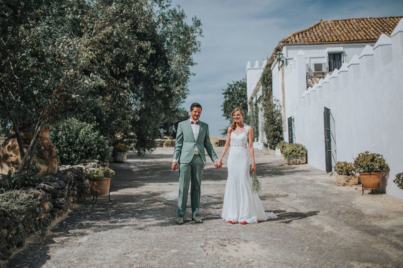 A couple walks hand in hand on a sunlit path with white Mediterranean buildings and olive trees in Portugal.