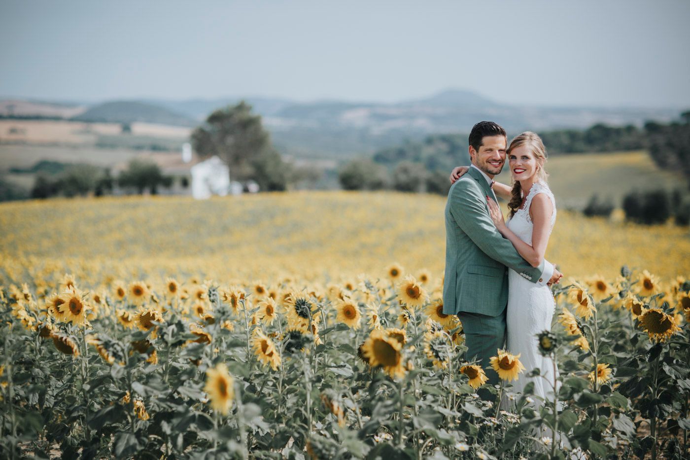 A couple stands together in a vibrant yellow sunflower field with mountains and a white chapel in the distance.