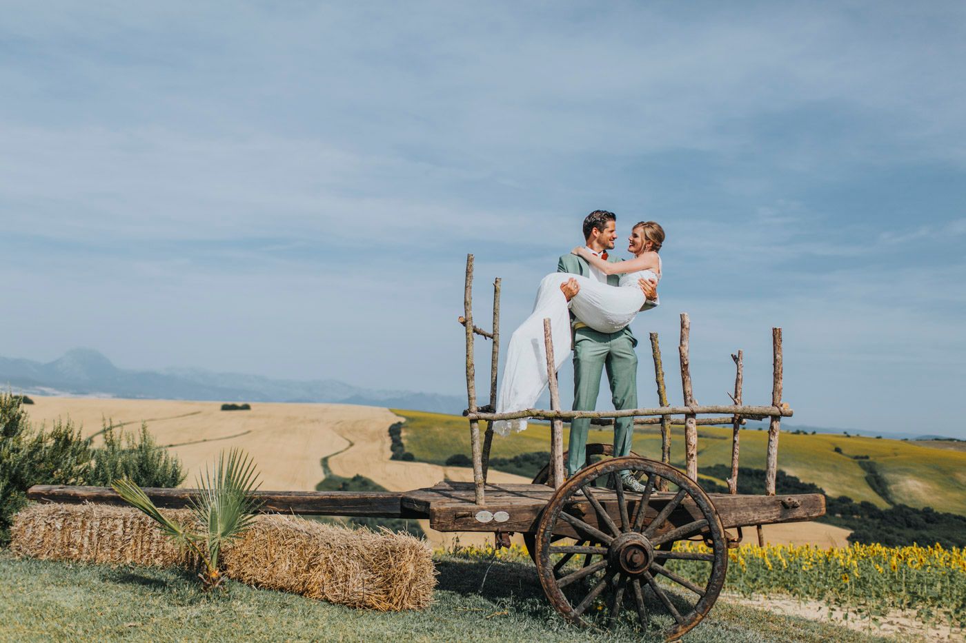 An elegant rustic scene with a couple standing on a vintage wooden cart in front of rolling golden hills.