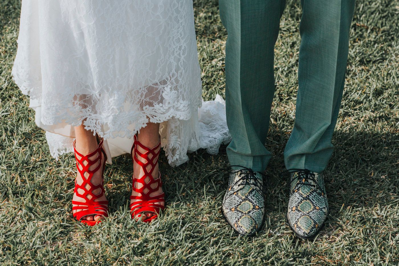 A bride in a white lace dress wearing bright red strappy heels stands next to someone in green pants with patterned loafers.
