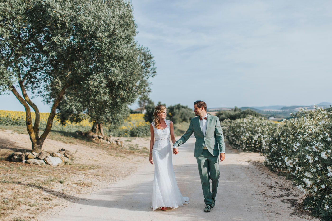 A couple walks hand in hand along a sunny Mediterranean path lined with olive trees on their wedding day.