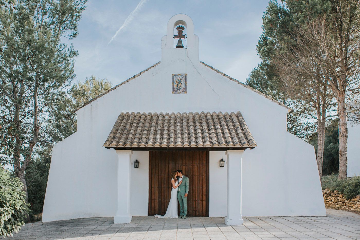 A white chapel with wooden doors and terracotta roof tiles stands nestled among pine trees on a sunny day.