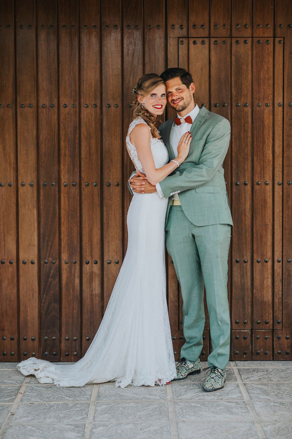 A bride in a flowing white gown and groom in a sage green suit pose together against a rustic wooden door background.