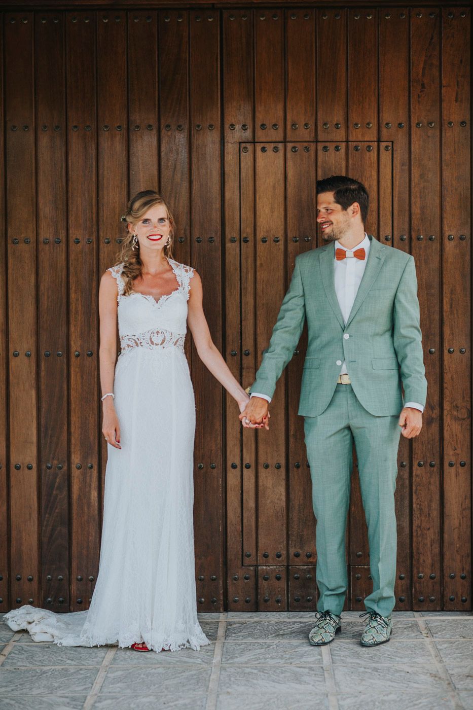 A bride in a white gown and groom in a mint green suit with red bowtie stand holding hands against wooden doors.