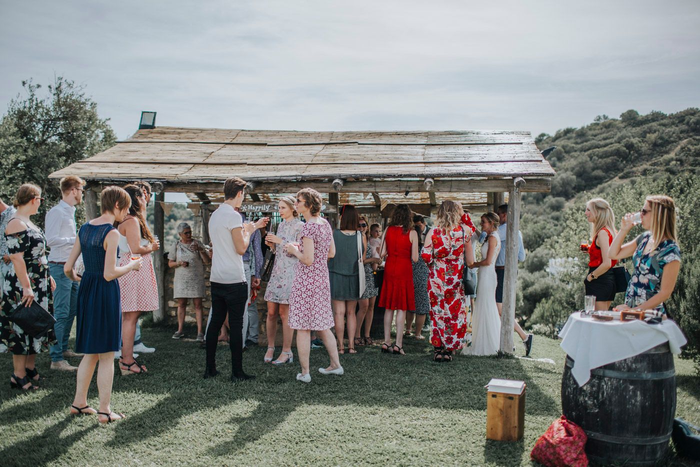 A group of wedding guests gather under a covered outdoor venue space during a daytime celebration.