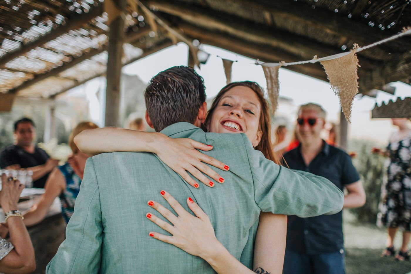 Two people embrace in a warm hug under a covered patio while guests look on in the background.
