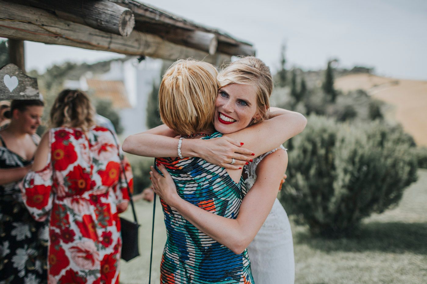People embrace at an outdoor event wearing colorful dresses against a natural landscape with rolling hills in the background.
