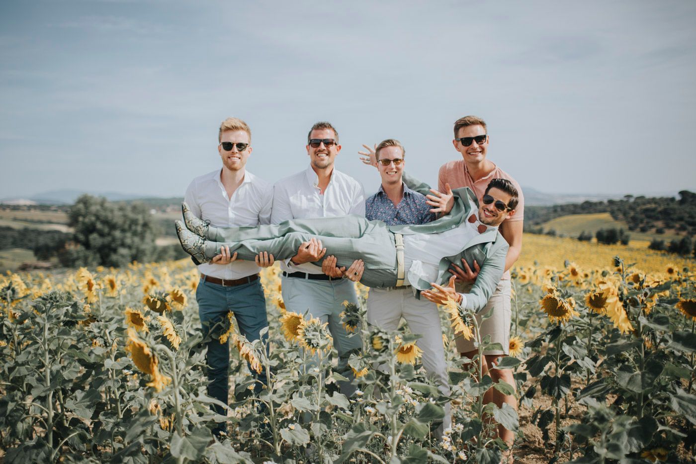 Group posing together in a vibrant sunflower field on a summer day with blue skies and rolling hills in background.