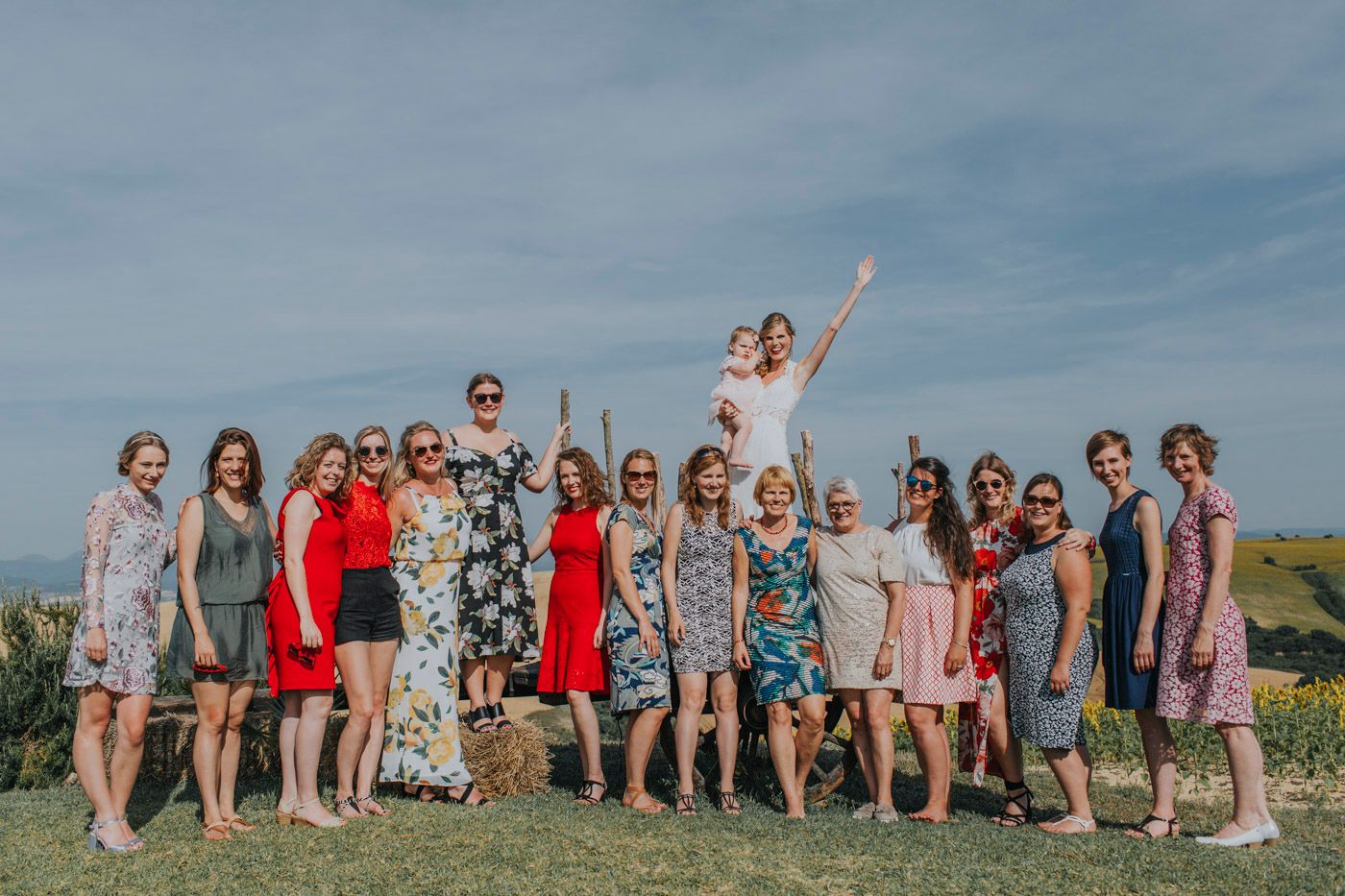 Large group in colorful summer dresses gathered outdoors for a celebratory photo against blue sky backdrop.