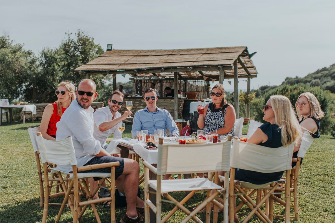 Group enjoying an outdoor meal at rustic wooden table with chairs beneath open-air structure in garden setting.
