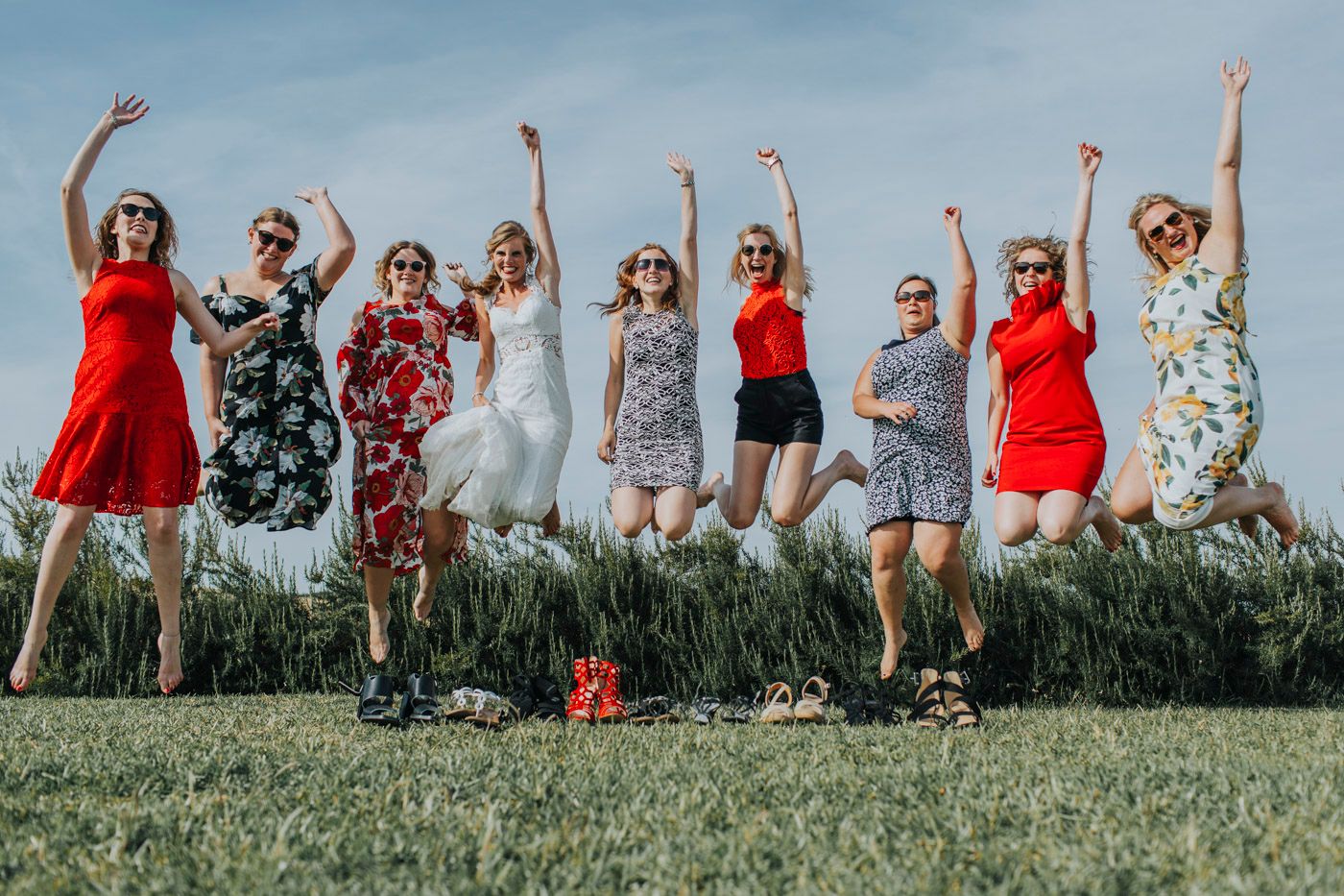 Group in colorful summer dresses jumping joyfully together on grassy field against blue sky.