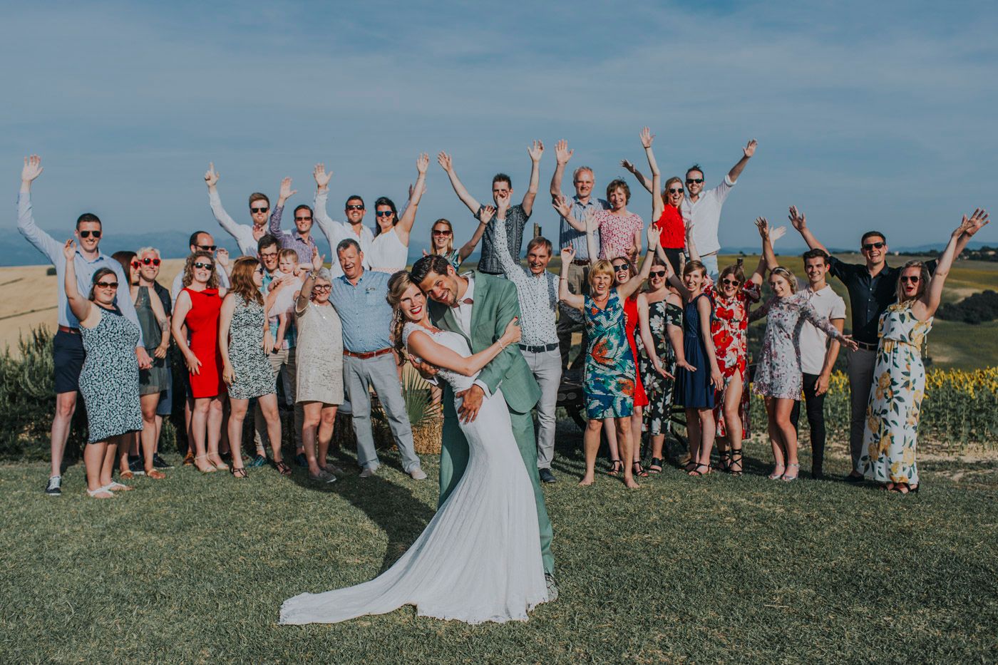 A celebratory group photo on a grassy hilltop with guests cheering and a couple posing in the center on their wedding day.