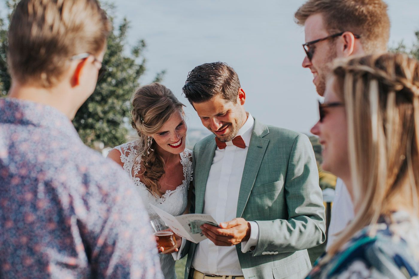 A joyful outdoor summer gathering shows people looking at something on paper while sharing laughs in natural sunlight.