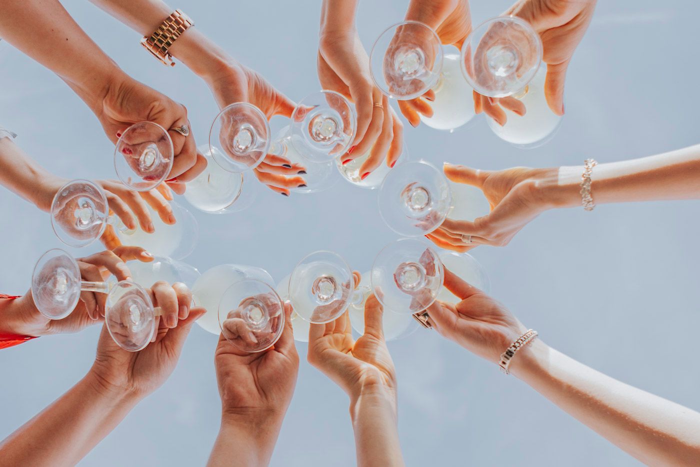 Multiple hands raise glasses in a celebratory circle toast from below against a bright sky background.