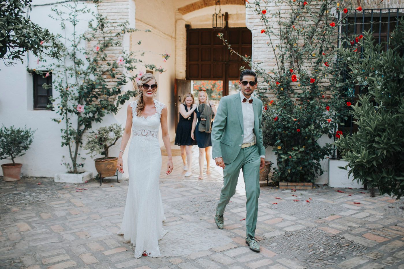 A couple in wedding attire walks along a stone path lined with blooming red flowers and white stucco walls.