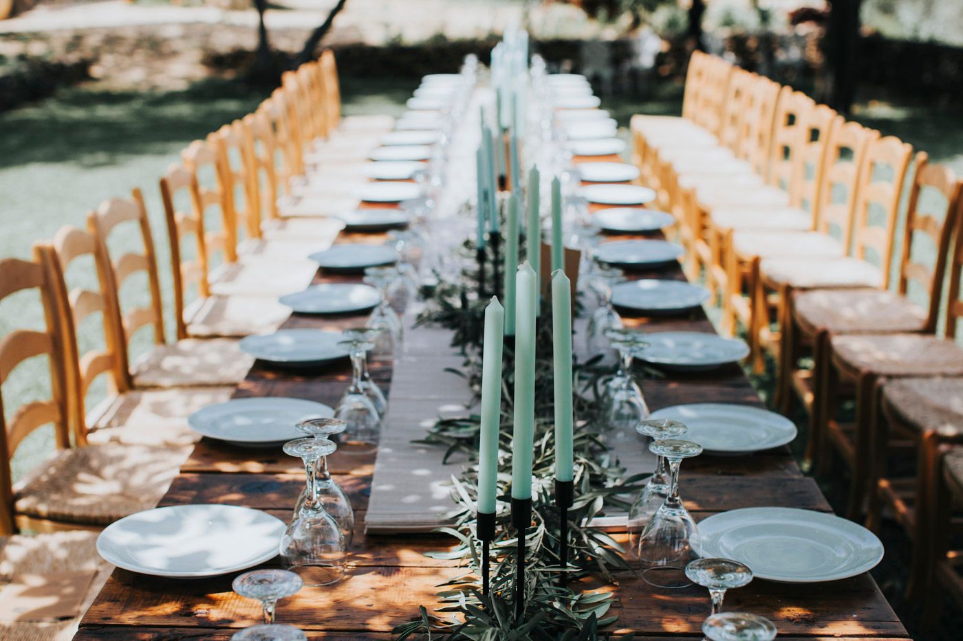 Long outdoor dining table set with gray plates, wooden chairs, and teal candles creating an elegant wedding reception setup.