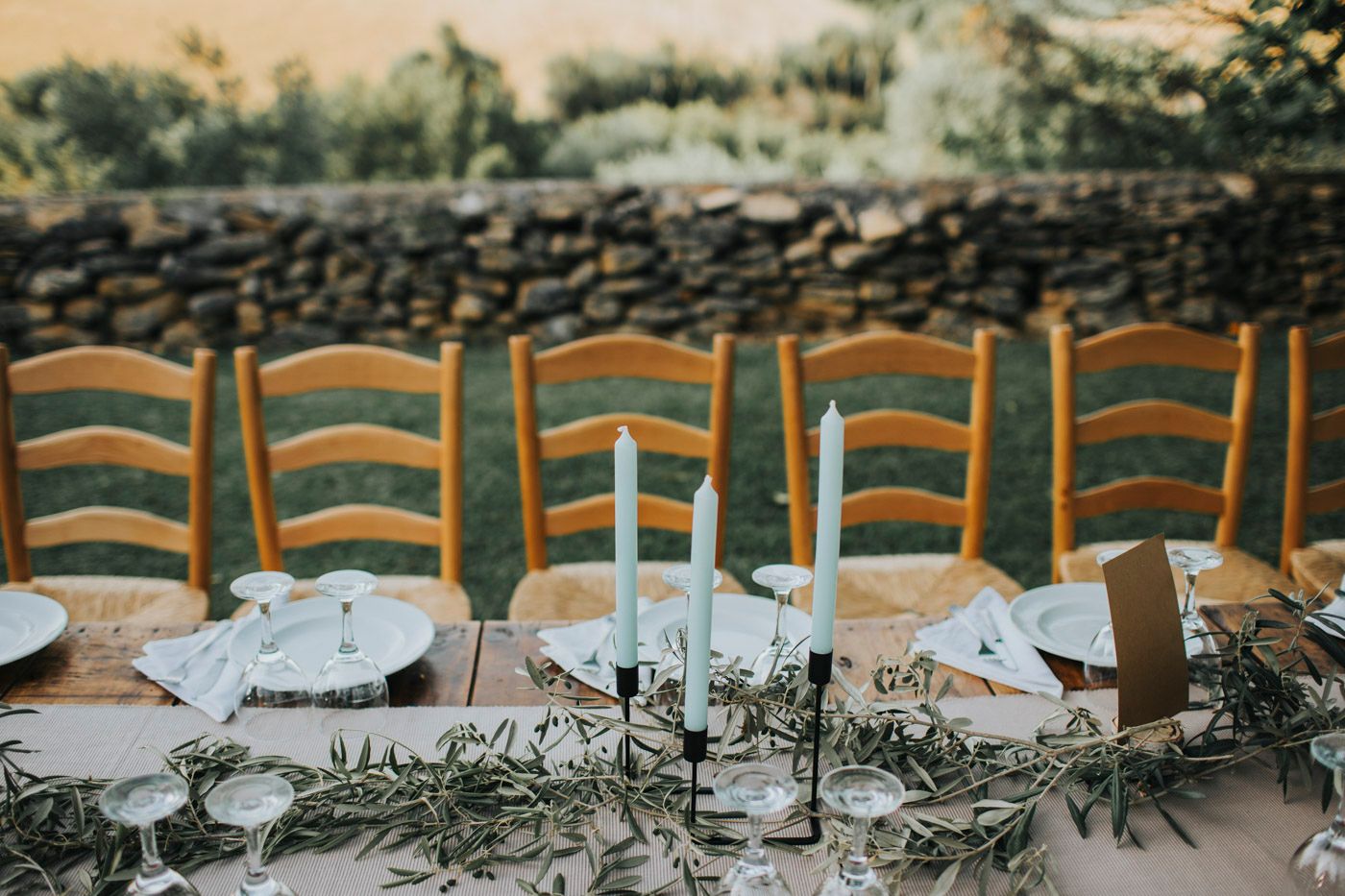 Rustic wooden chairs line an elegant outdoor table setting decorated with delicate greenery and white place settings.