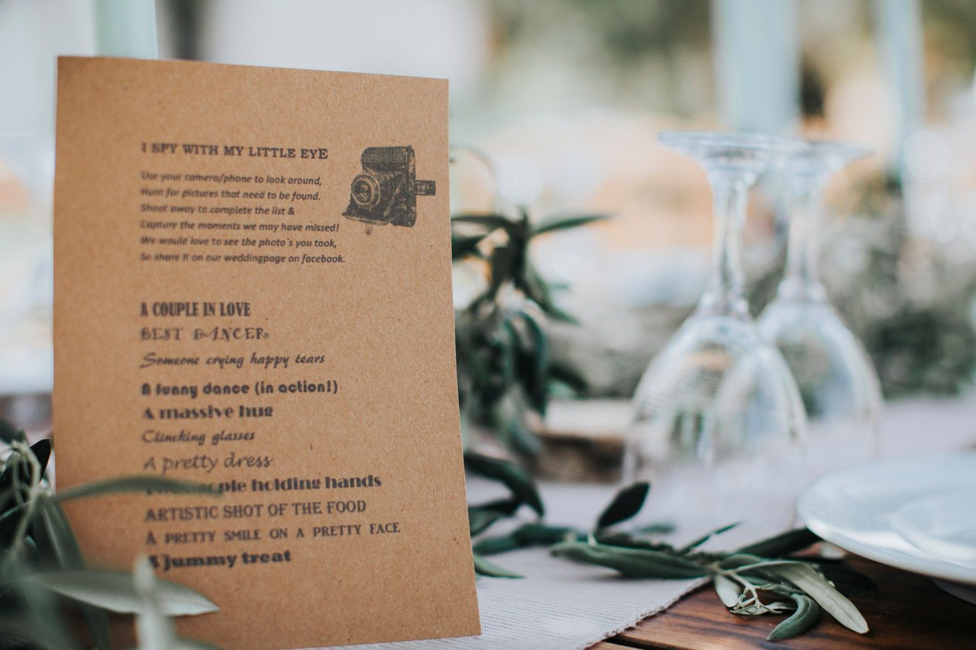 A brown kraft paper menu card sits on a table with wine glasses and greenery in soft focus background.