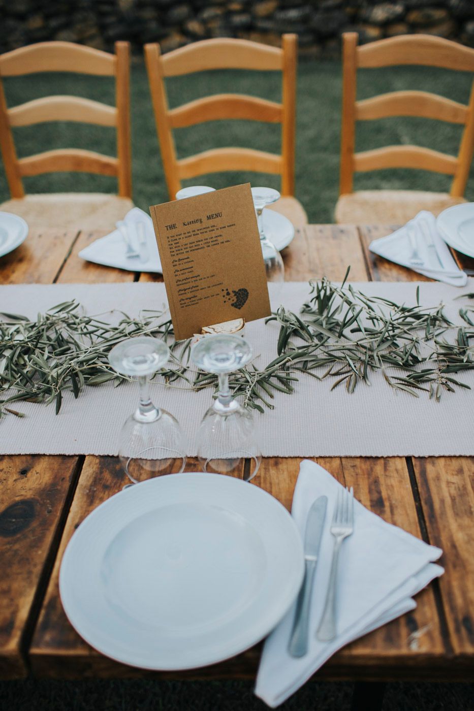 Rustic table setting with white plates, glasses and greenery centerpiece featuring a kraft paper menu on a wooden surface.