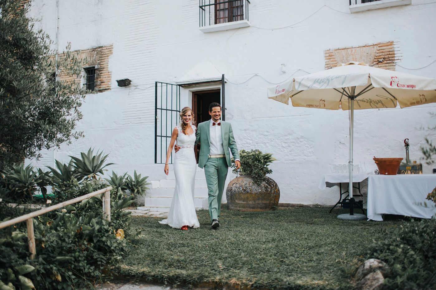 A couple in wedding attire walks together in a garden courtyard with white walls and a beige umbrella.