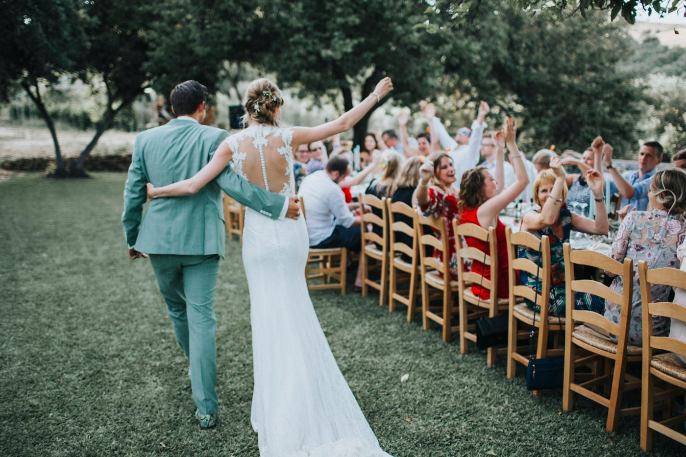 Wedding guests seated in wooden chairs cheer and wave while couple walks down outdoor aisle at garden ceremony.