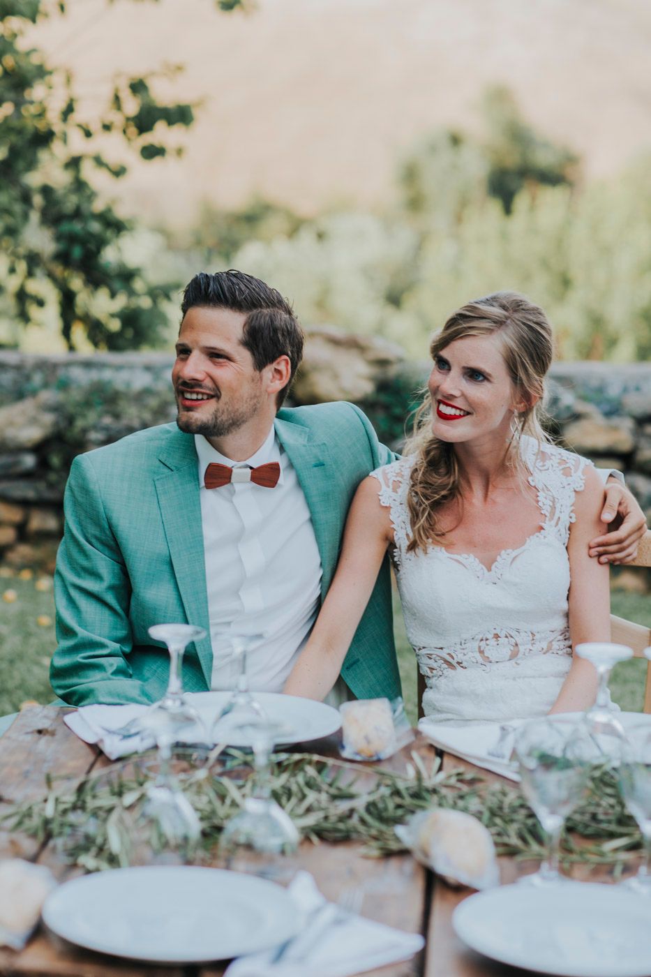 A happy couple sits together at their rustic outdoor wedding reception wearing a mint green suit and lace wedding dress.