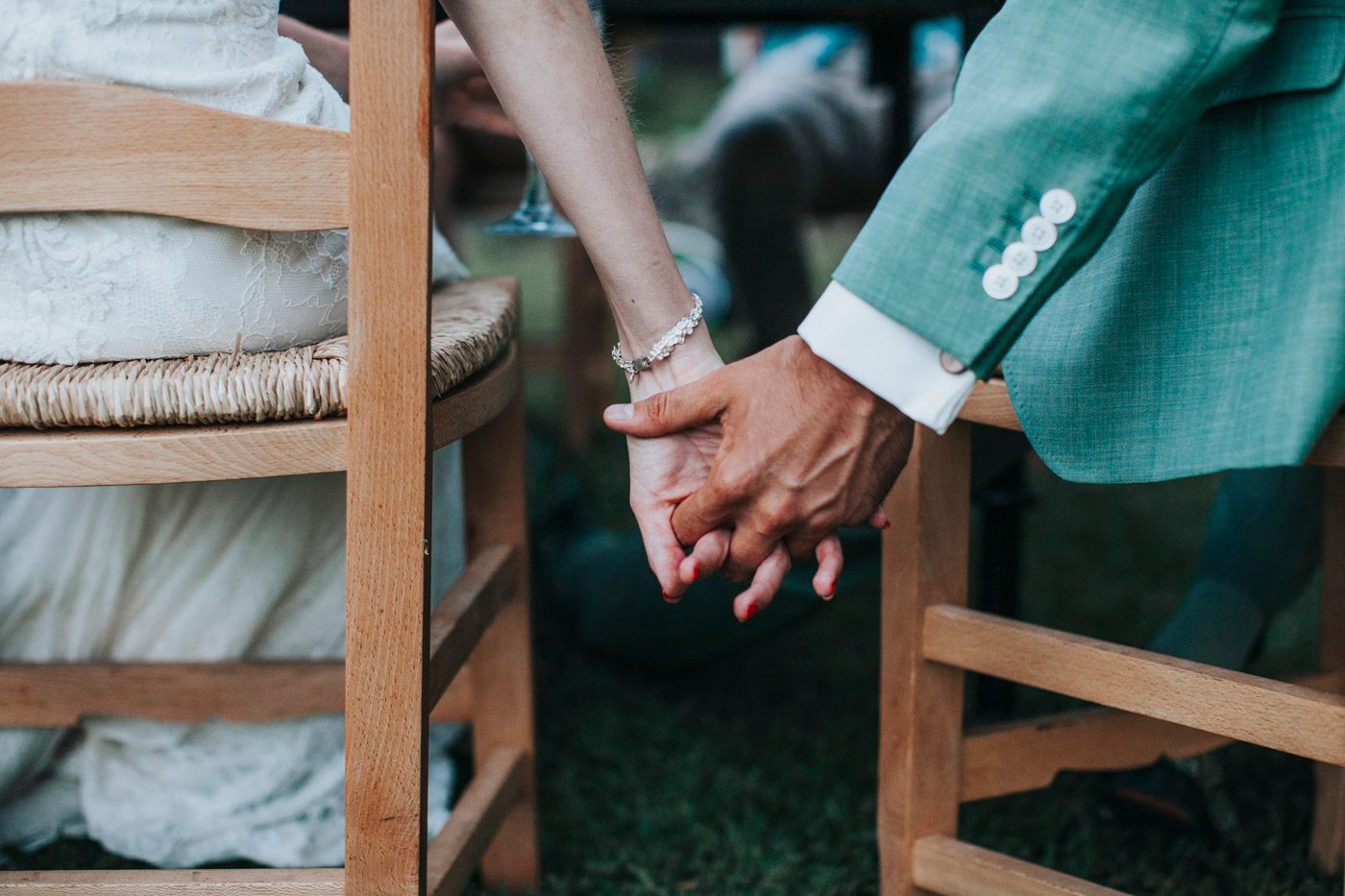 Close-up view of two people holding hands while sitting in wooden chairs on grass during an outdoor event.
