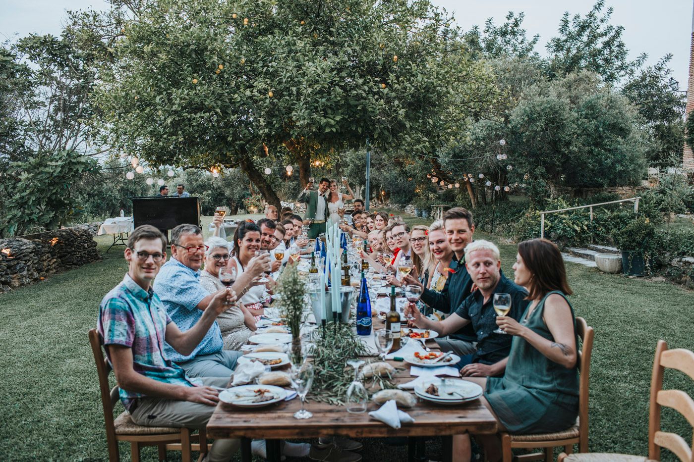 Large group dining at rustic outdoor table setting with blue table runner and natural garden backdrop.