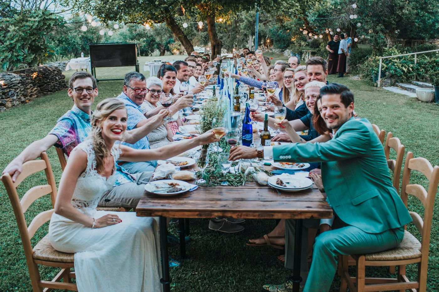 Wedding guests seated at a long outdoor table raise glasses in a celebratory toast during a garden reception.