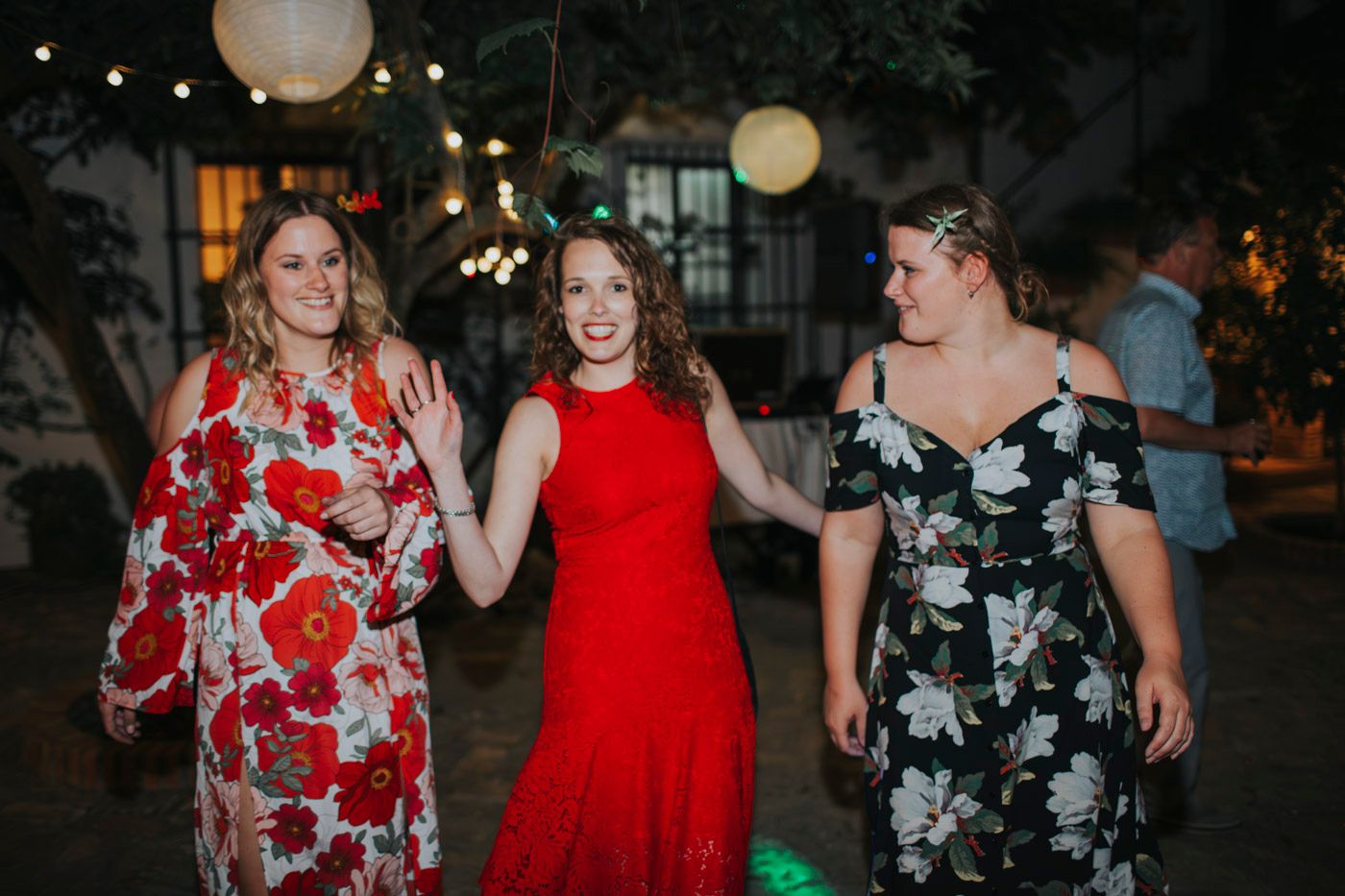 Three guests in colorful dresses dancing together at an evening outdoor celebration with lanterns overhead.