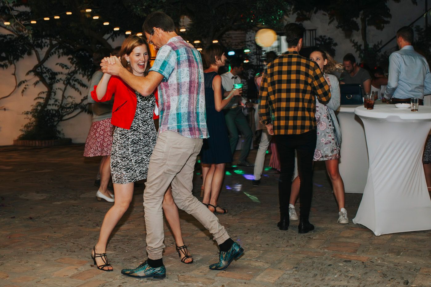 Couples dance together at an outdoor evening party with string lights overhead and cocktail tables in the background.