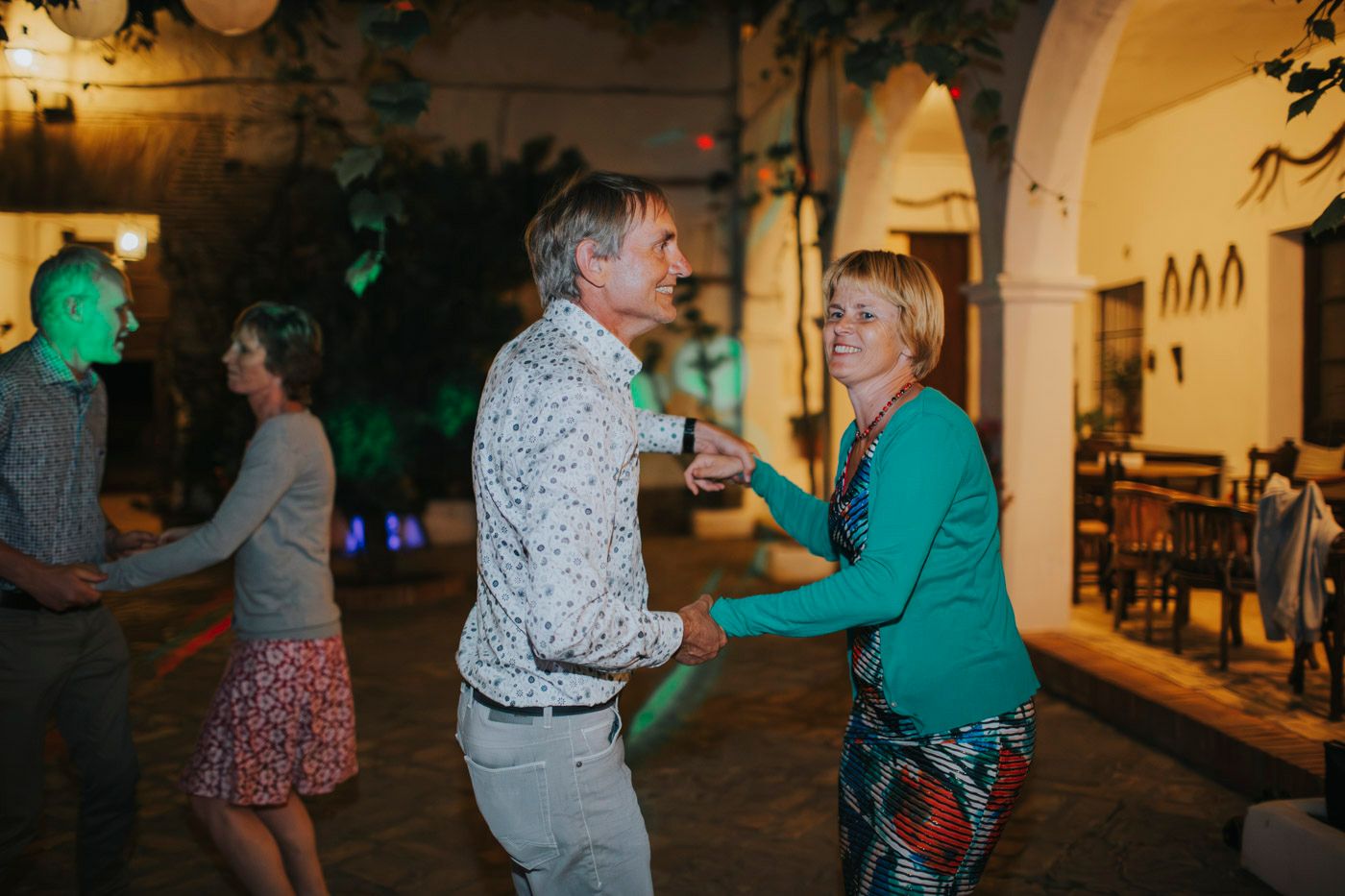 A couple in green and patterned shirts dance together at an evening event under arched doorways.